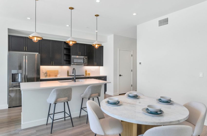 Modern kitchen with dark cabinets, white countertops, and a marble dining table set for four, illuminated by pendant lights.