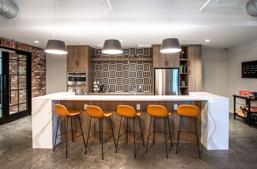 Modern office kitchenette with wooden cabinets, stainless steel appliances, and a white marble bar with orange stools.