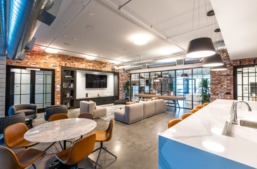 Modern office lounge with stylish furniture, exposed brick walls, wood floors, pendant lighting, and a large tv.