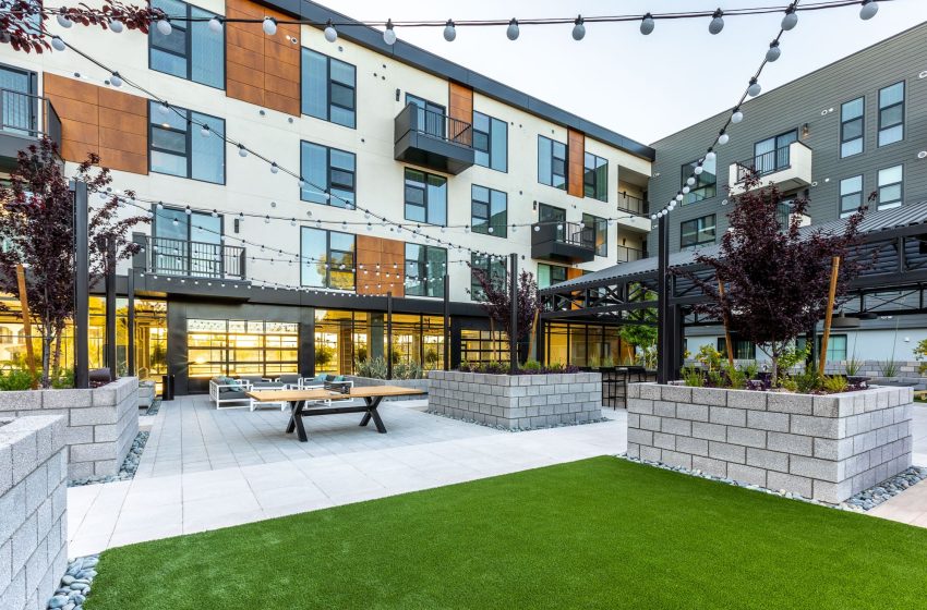 Modern apartment building courtyard with green artificial grass, string lights, picnic tables, and surrounding residential units with large glass windows.
